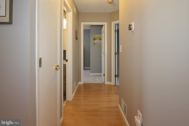 hallway featuring light wood finished floors, visible vents, a textured ceiling, and baseboards