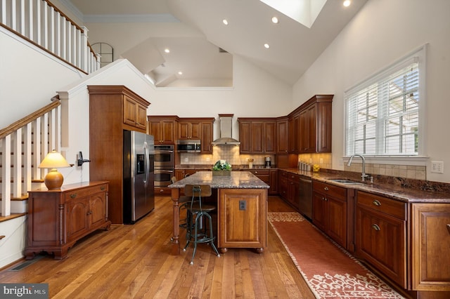 kitchen with a sink, stainless steel appliances, wall chimney exhaust hood, light wood-type flooring, and a center island