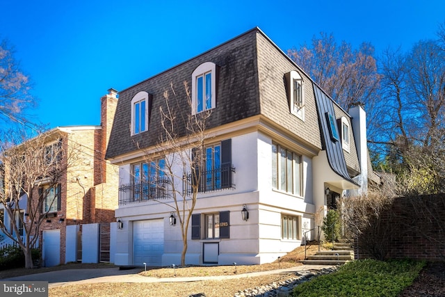 exterior space with stucco siding, mansard roof, an attached garage, a shingled roof, and a chimney