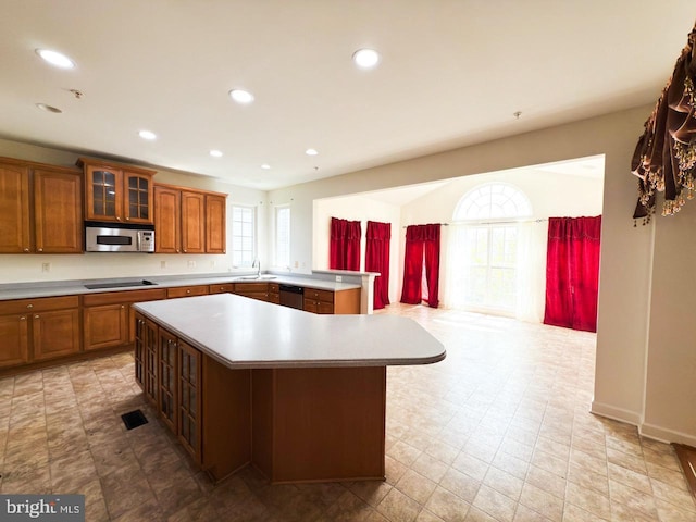 kitchen with stainless steel appliances, a center island, glass insert cabinets, brown cabinetry, and light countertops