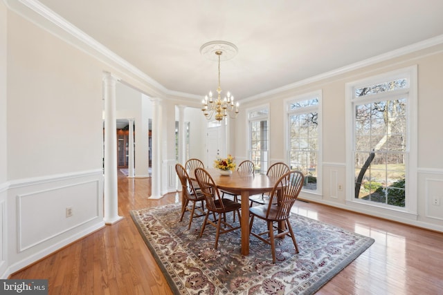 dining area with a notable chandelier, wainscoting, light wood finished floors, crown molding, and ornate columns