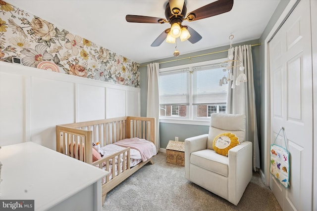 carpeted bedroom with a wainscoted wall, a crib, a closet, wallpapered walls, and ceiling fan