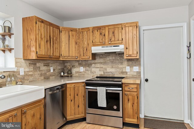 kitchen featuring tasteful backsplash, under cabinet range hood, light countertops, brown cabinets, and appliances with stainless steel finishes