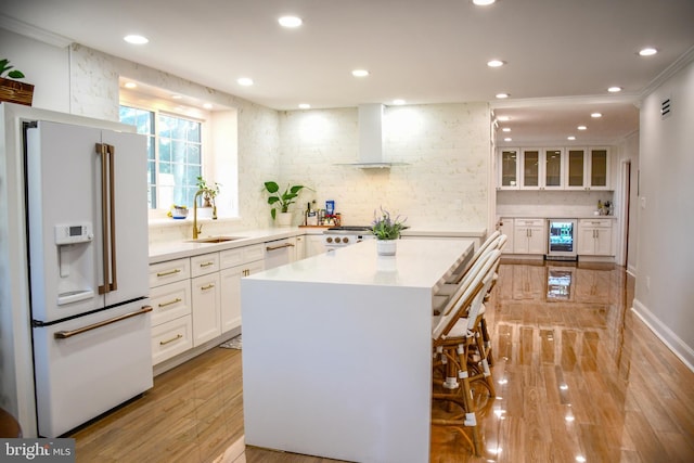 kitchen with wall chimney range hood, wine cooler, white appliances, white cabinetry, and a sink