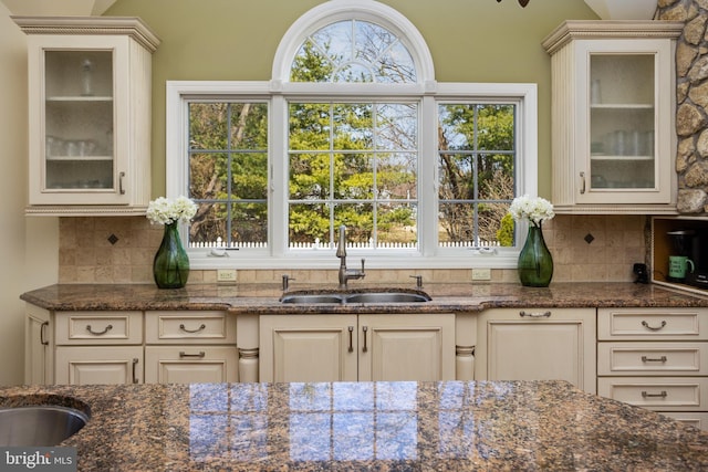 kitchen with backsplash, dark stone counters, glass insert cabinets, and a sink