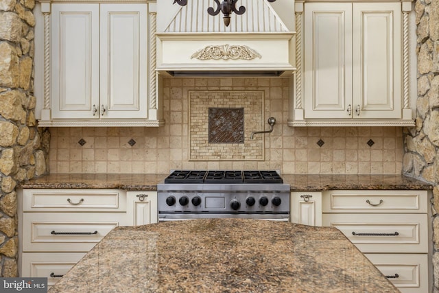 kitchen with dark stone countertops, stove, custom range hood, cream cabinetry, and backsplash