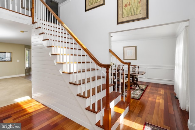 staircase with a decorative wall, wood finished floors, a towering ceiling, and ornamental molding