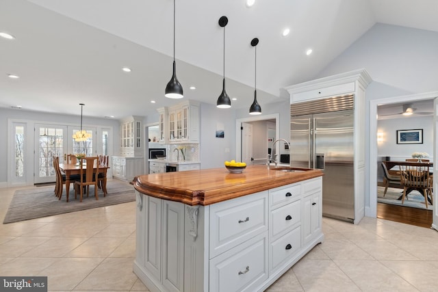 kitchen with stainless steel built in refrigerator, a center island with sink, a sink, light tile patterned floors, and glass insert cabinets