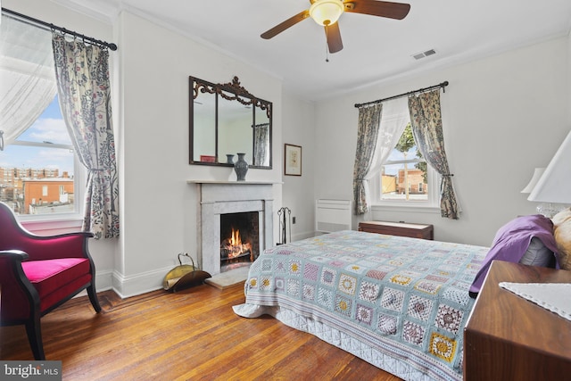 bedroom with visible vents, a warm lit fireplace, wood finished floors, baseboards, and ceiling fan