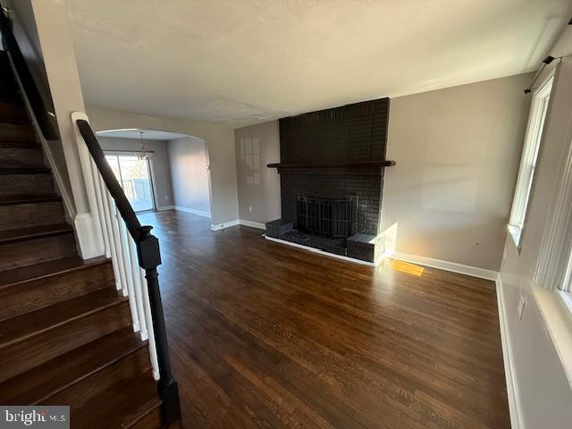 unfurnished living room with stairway, arched walkways, a fireplace, baseboards, and dark wood-style flooring