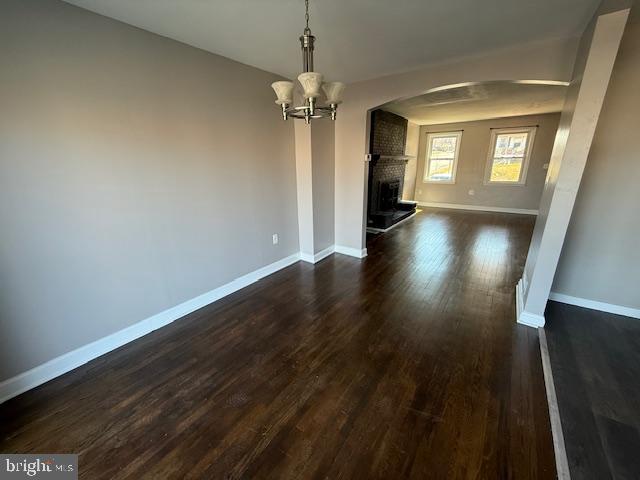 unfurnished dining area with arched walkways, an inviting chandelier, a fireplace, baseboards, and dark wood-style flooring