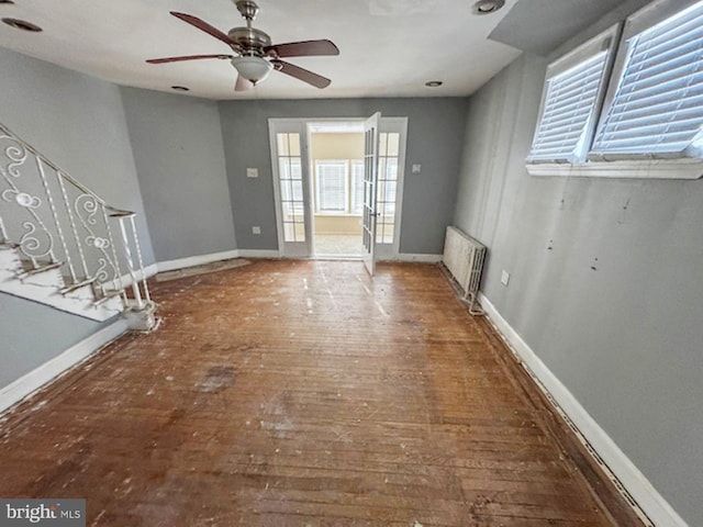 unfurnished living room featuring hardwood / wood-style flooring, radiator heating unit, stairway, baseboards, and ceiling fan