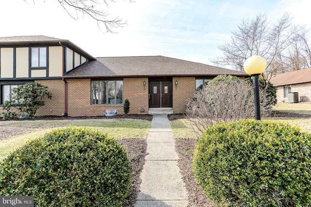 tudor-style house with cooling unit, stucco siding, a shingled roof, a front lawn, and brick siding