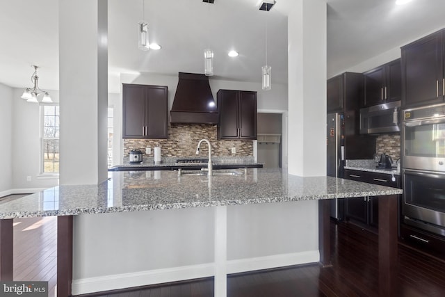 kitchen with a sink, premium range hood, light stone counters, appliances with stainless steel finishes, and a kitchen island with sink