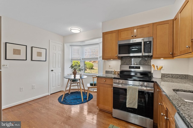 kitchen featuring light wood-style flooring, stone countertops, appliances with stainless steel finishes, brown cabinetry, and baseboards