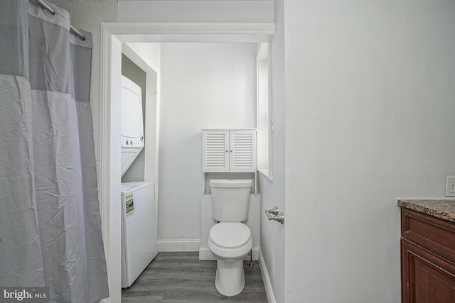 bathroom with toilet, stacked washer and clothes dryer, wood finished floors, baseboards, and vanity