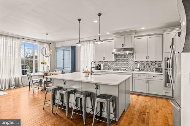 kitchen featuring a sink, light countertops, under cabinet range hood, a kitchen bar, and backsplash