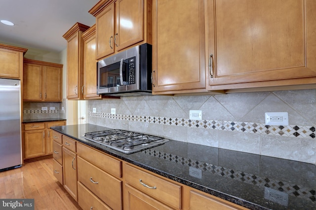 kitchen featuring light wood-type flooring, backsplash, dark stone counters, appliances with stainless steel finishes, and brown cabinetry