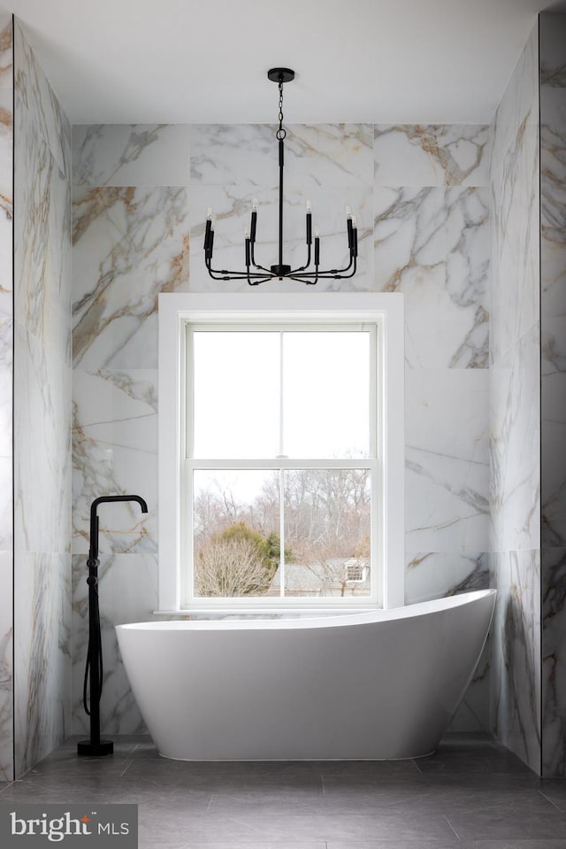 bathroom featuring a notable chandelier, a freestanding bath, and stone wall