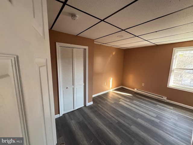 unfurnished bedroom with baseboards, dark wood finished floors, a drop ceiling, a closet, and a baseboard radiator