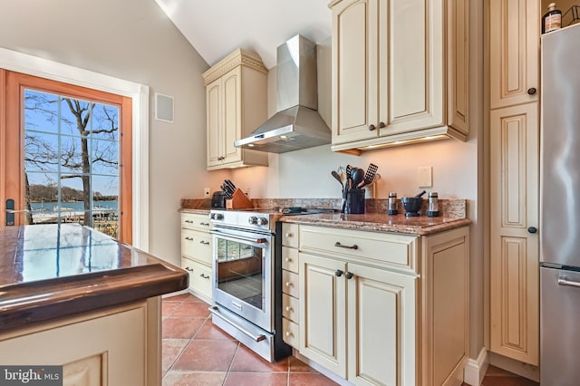 kitchen with wall chimney range hood, vaulted ceiling, light tile patterned floors, cream cabinets, and stainless steel appliances