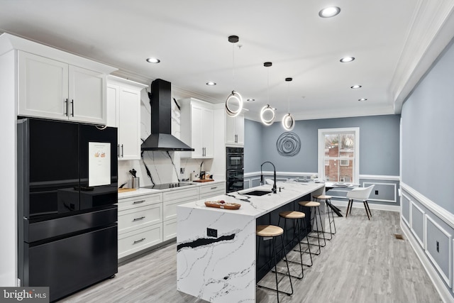 kitchen with a wainscoted wall, black appliances, a center island with sink, white cabinetry, and wall chimney range hood