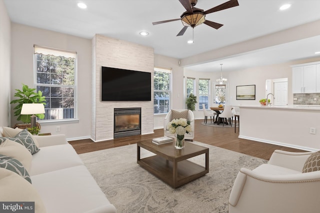 living area featuring baseboards, recessed lighting, a fireplace, ceiling fan with notable chandelier, and dark wood-style floors