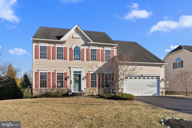 colonial inspired home featuring driveway, stone siding, roof with shingles, a front yard, and an attached garage