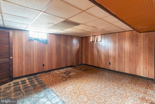 basement with tile patterned floors, wooden walls, baseboards, and a drop ceiling