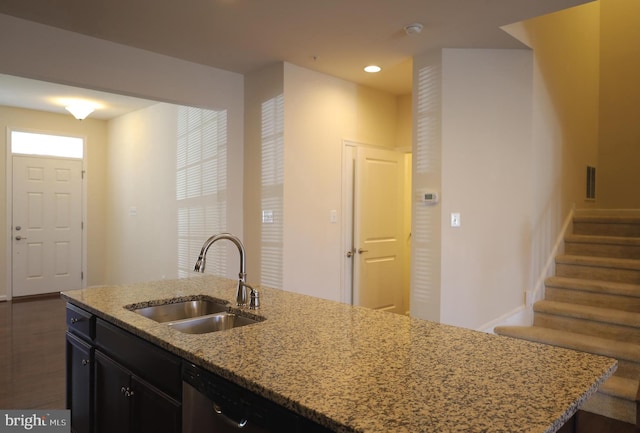 kitchen featuring light stone counters, an island with sink, a sink, dishwasher, and dark cabinets