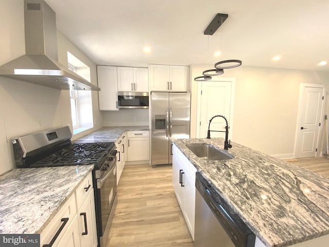 kitchen featuring a kitchen island with sink, a sink, stainless steel appliances, white cabinets, and wall chimney range hood