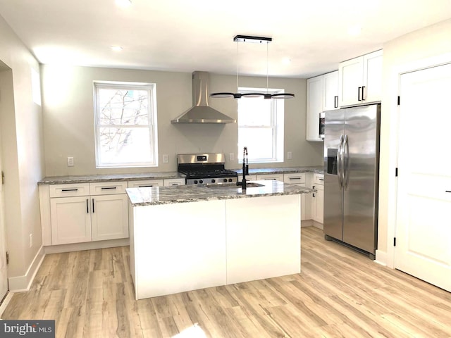 kitchen featuring wall chimney range hood, a center island with sink, light stone counters, appliances with stainless steel finishes, and light wood-style floors