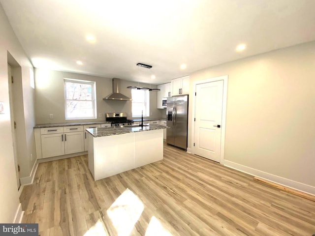 kitchen with wall chimney range hood, an island with sink, light wood-style flooring, white cabinets, and stainless steel appliances