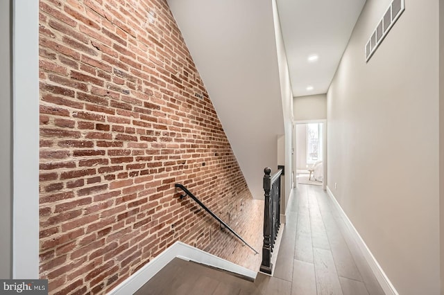 hall with wood finished floors, visible vents, brick wall, baseboards, and an upstairs landing