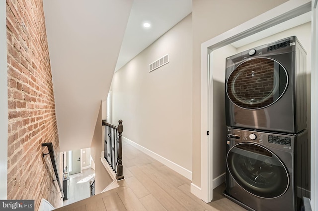 laundry area featuring stacked washer / dryer, visible vents, brick wall, baseboards, and wood finished floors