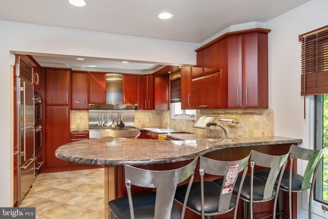 kitchen featuring reddish brown cabinets, a peninsula, wall chimney exhaust hood, and stainless steel oven