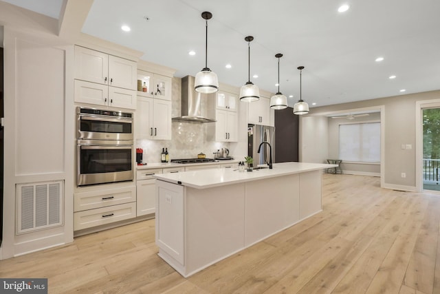 kitchen featuring visible vents, wall chimney range hood, light wood-style flooring, appliances with stainless steel finishes, and a kitchen island with sink