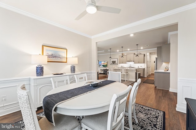 dining area featuring ceiling fan, a wainscoted wall, ornamental molding, dark wood-style floors, and a decorative wall