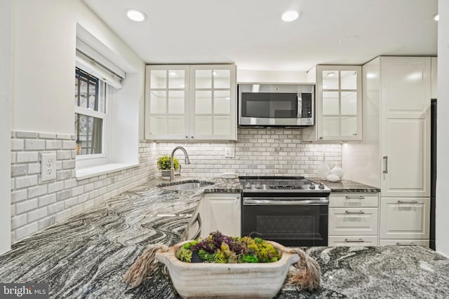 kitchen with glass insert cabinets, dark stone countertops, appliances with stainless steel finishes, white cabinets, and a sink