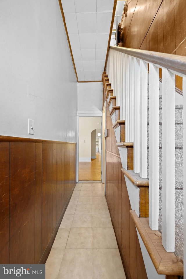 corridor featuring light tile patterned floors, arched walkways, stairs, wood walls, and wainscoting