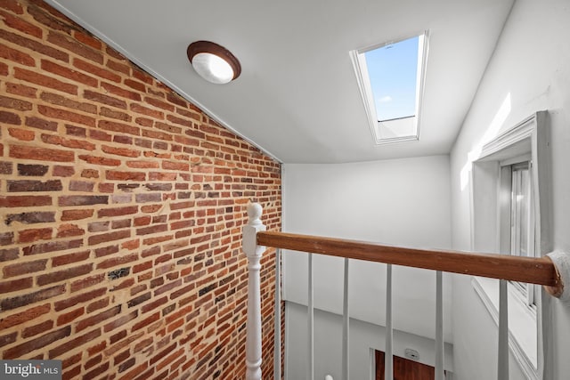 hallway featuring lofted ceiling with skylight, an upstairs landing, and brick wall