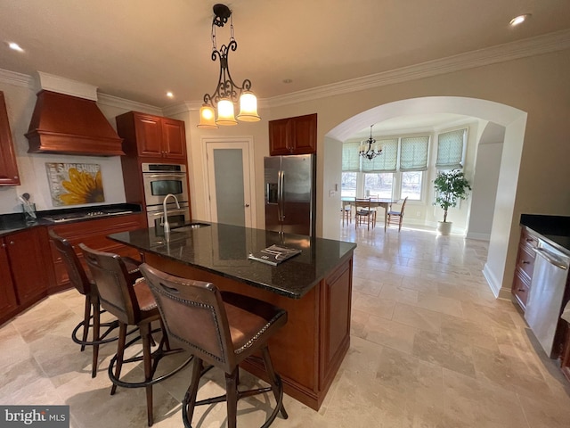 kitchen featuring custom exhaust hood, an island with sink, arched walkways, stainless steel appliances, and a notable chandelier