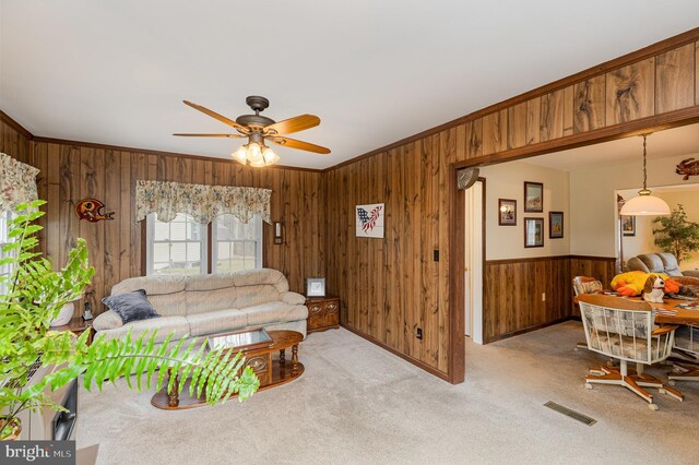 living room featuring carpet flooring, visible vents, wood walls, ornamental molding, and a ceiling fan