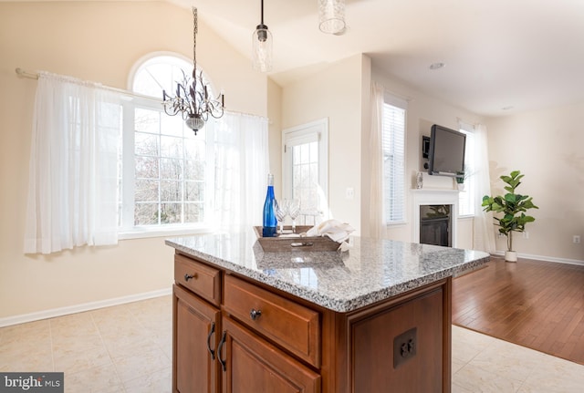 kitchen featuring brown cabinetry, light stone countertops, a fireplace, hanging light fixtures, and vaulted ceiling