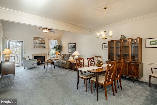 dining space featuring crown molding, a warm lit fireplace, ceiling fan with notable chandelier, and carpet floors