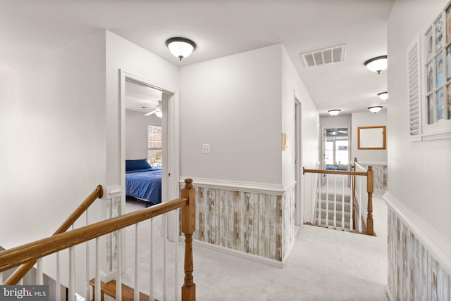 hallway featuring a wainscoted wall, visible vents, carpet floors, wood walls, and an upstairs landing