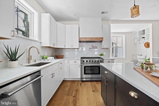 kitchen with light wood-type flooring, custom range hood, appliances with stainless steel finishes, white cabinets, and a sink