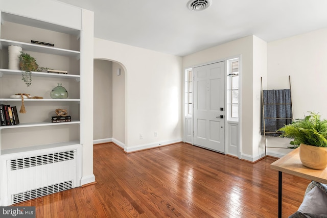 foyer entrance with visible vents, hardwood / wood-style flooring, radiator heating unit, arched walkways, and baseboards