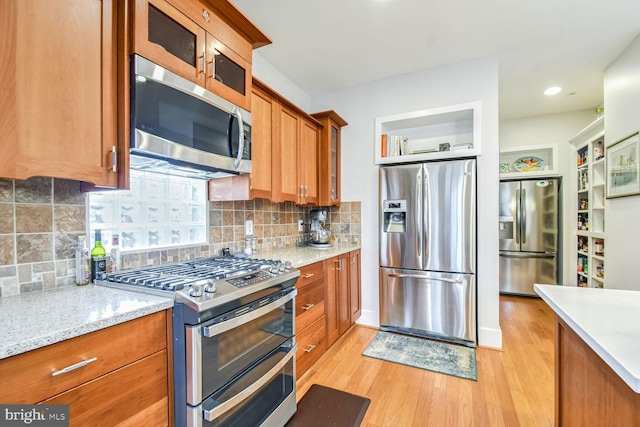 kitchen with backsplash, appliances with stainless steel finishes, brown cabinetry, light wood finished floors, and glass insert cabinets