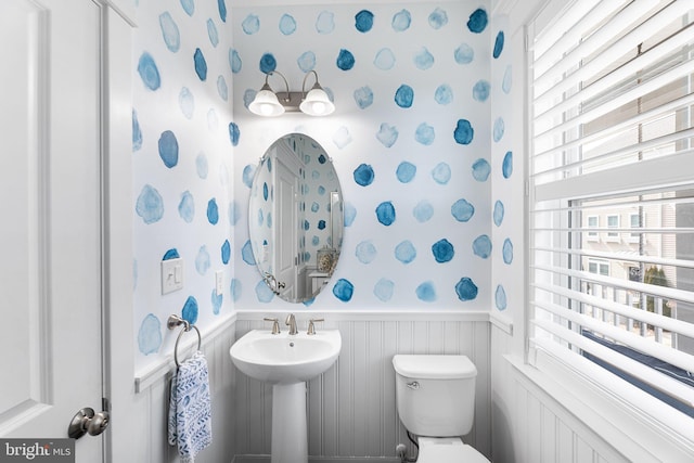 half bath with wallpapered walls, toilet, a sink, and wainscoting